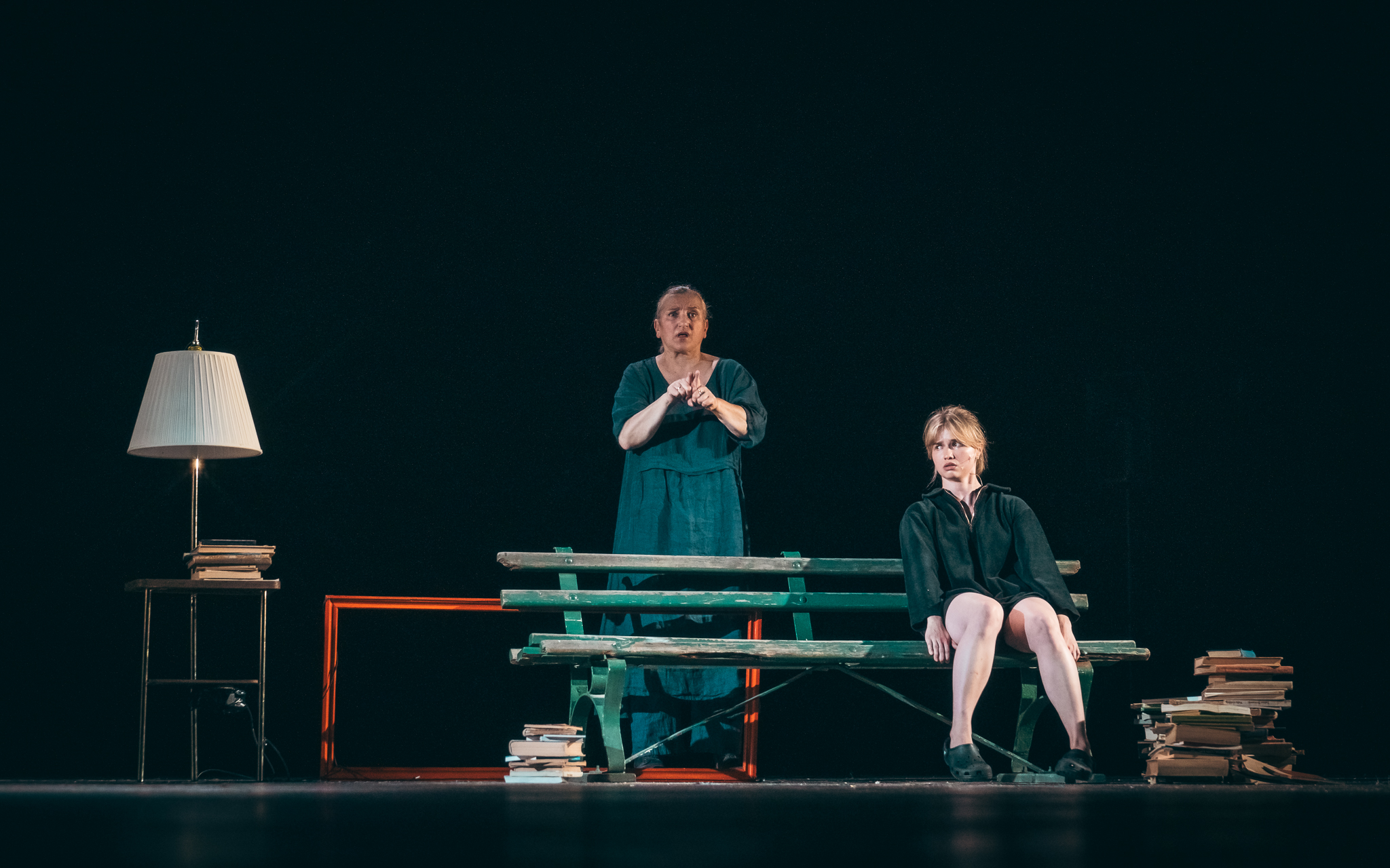 Na scenie znajdują się dwie osoby – jedna stoi i wykonuje gesty przypominające język migowy, a druga siedzi na ławce obok stosów książek, w otoczeniu lampy i minimalistycznej scenografii, w nastroju skupienia i ciszy.