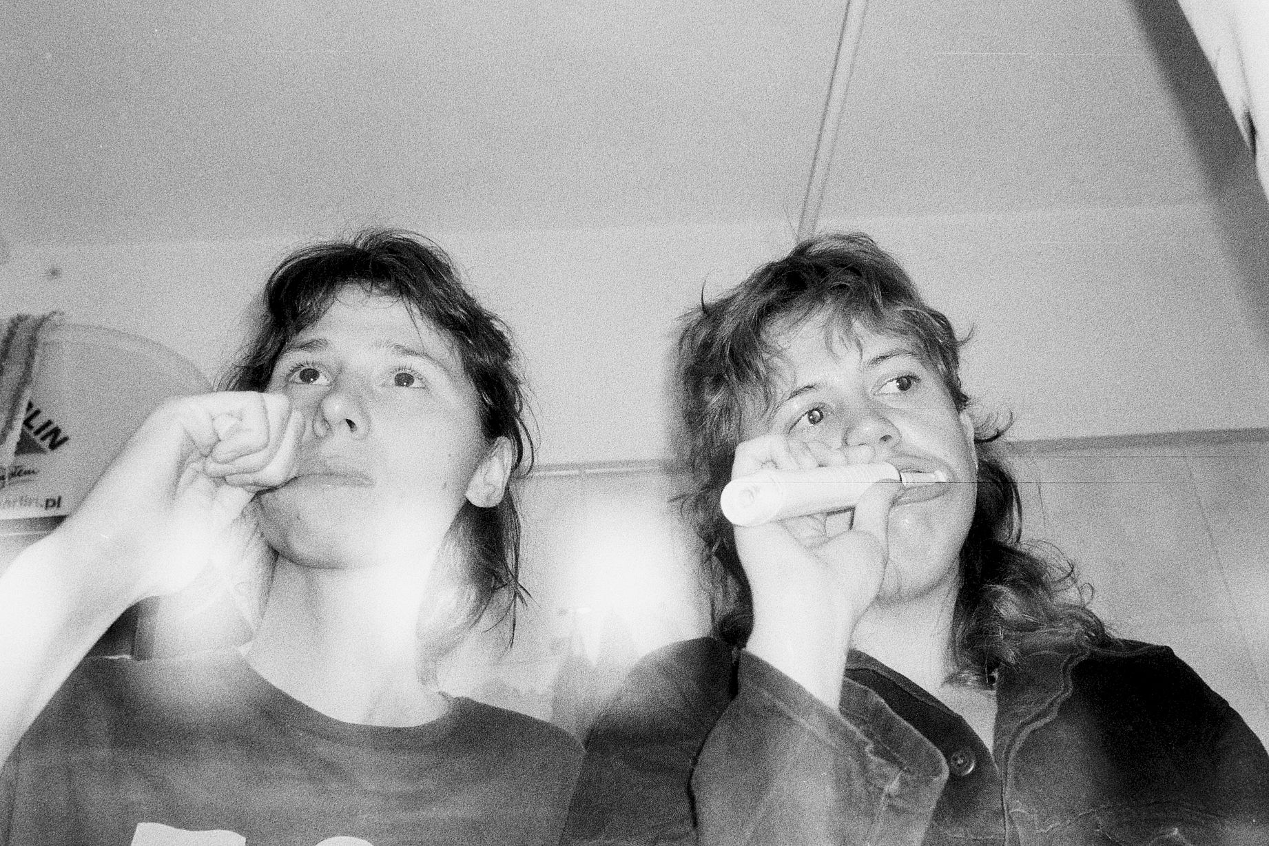 Czarno-białe, ziarniste zdjęcie ukazuje dwie osoby w ciasnym pomieszczeniu, z których jedna trzyma przy ustach podłużny, jasny przedmiot.