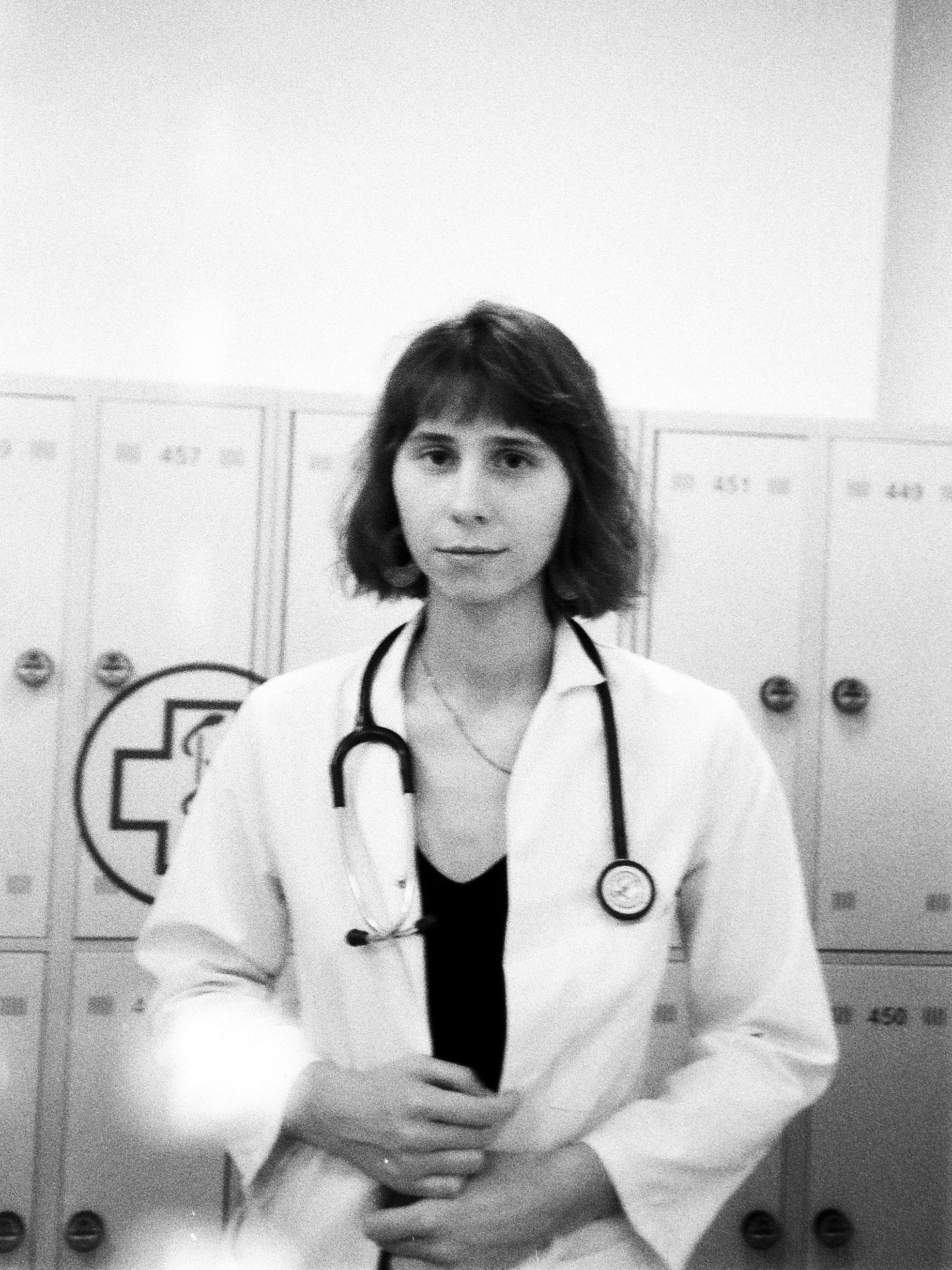 Czarno-białe zdjęcie przedstawia młodą kobietę w białym kitlu lekarskim ze stetoskopem na szyi, stojącą na tle szafek z symbolem medycznym.