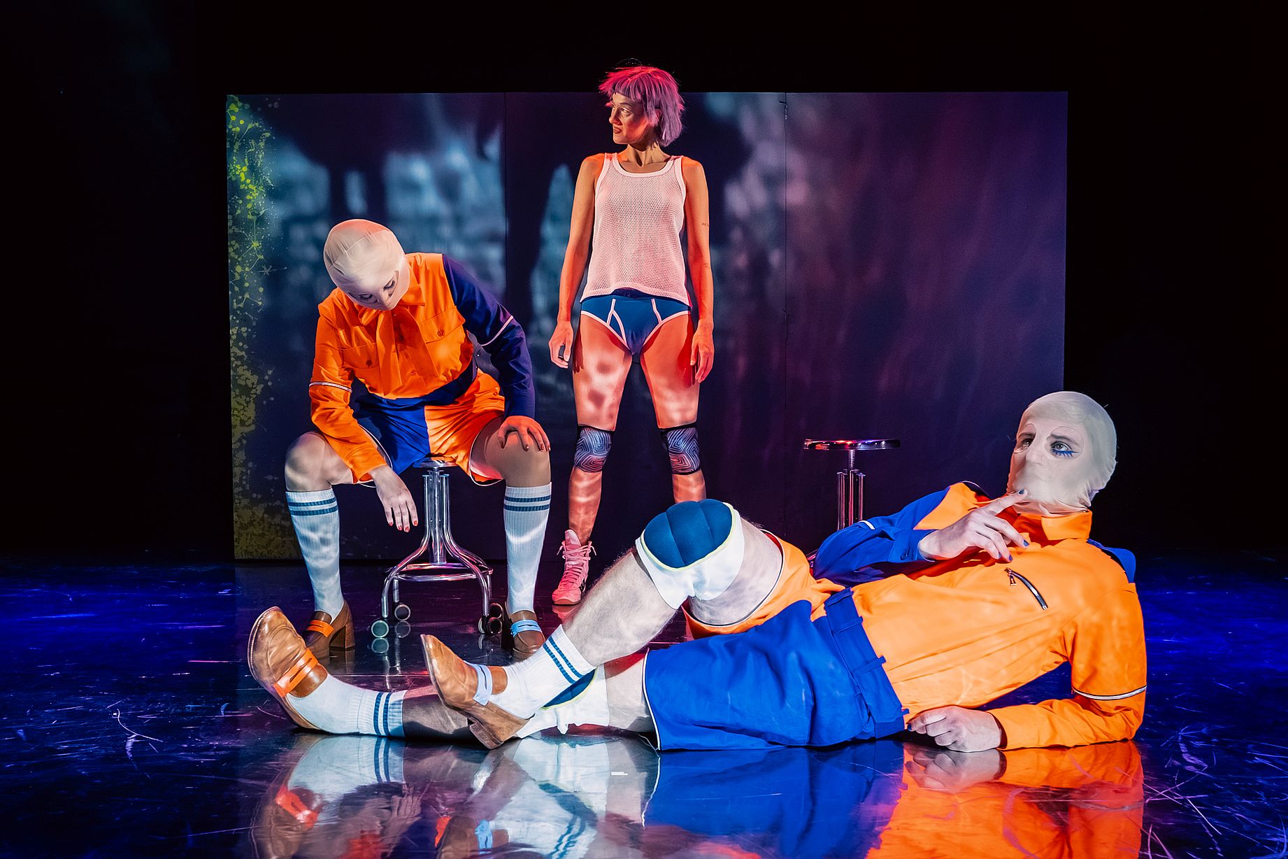 Scena w teatrze, dziwne stroje, pomarańczowe bluzy, niebieskie spodenki, dwóch aktorów w maskach, aktorka w majtkach