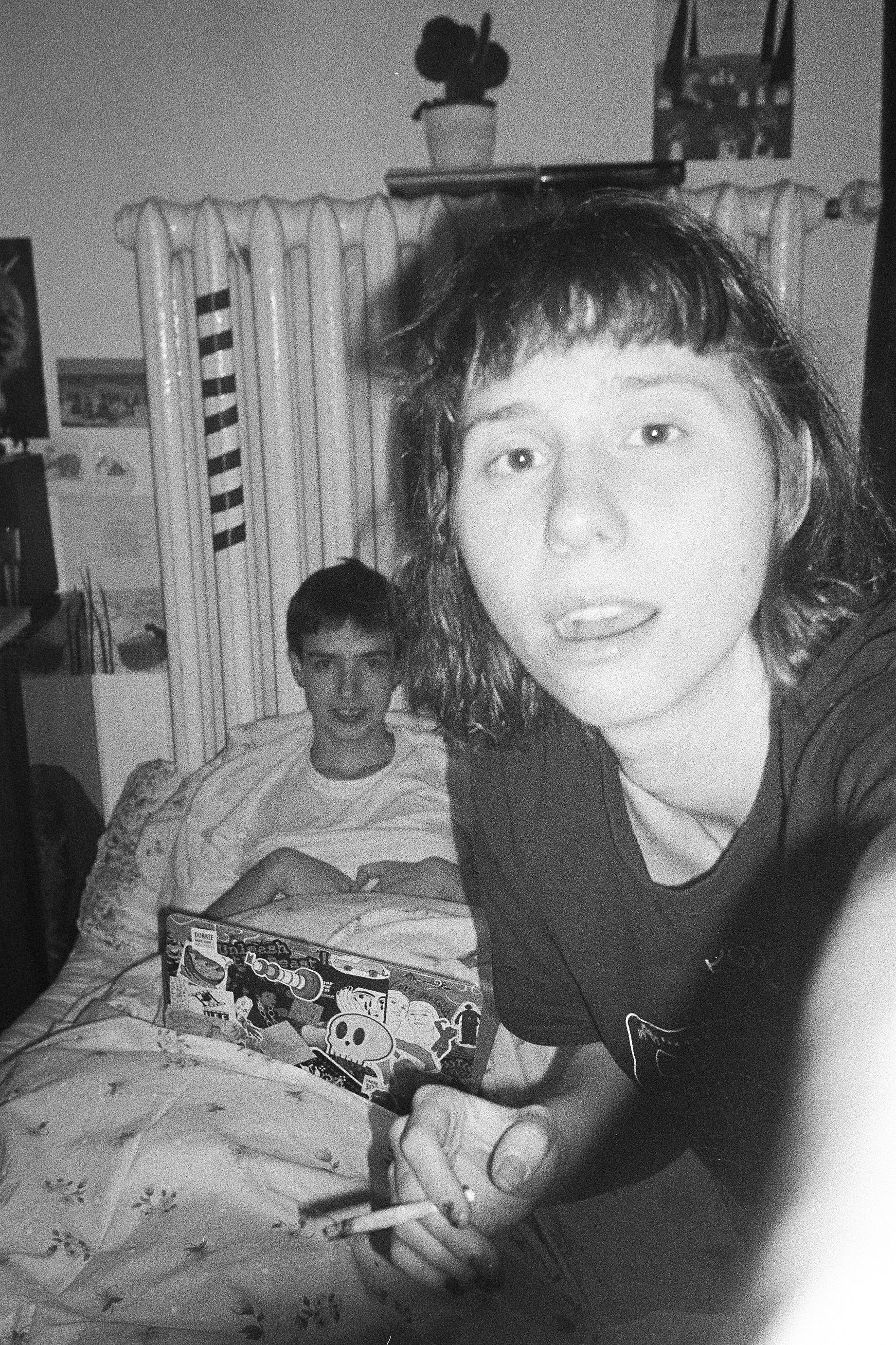 Czarno-białe, ziarniste zdjęcie przedstawia dwoje młodych ludzi w sypialni, z których kobieta na pierwszym planie trzyma papierosa, a mężczyzna w tle siedzi w łóżku.