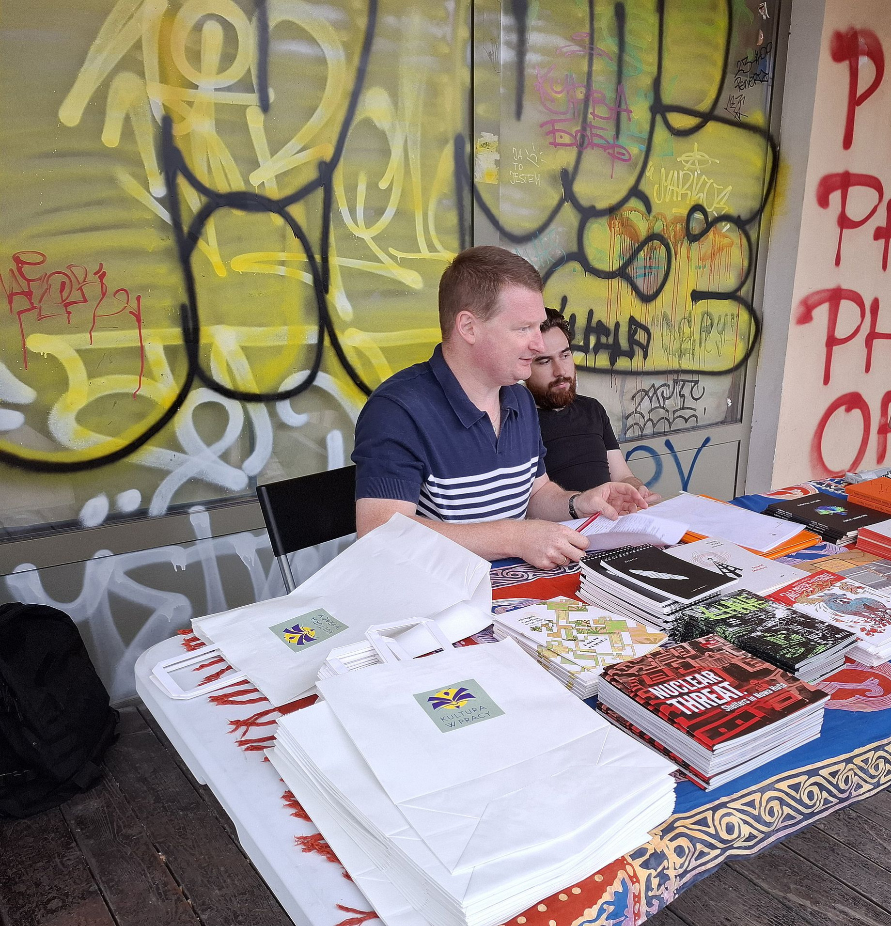 Pisanie wierszy na zamówienie na Placu Targowym, dwóch facetów siedzi przy stole pełnem książek