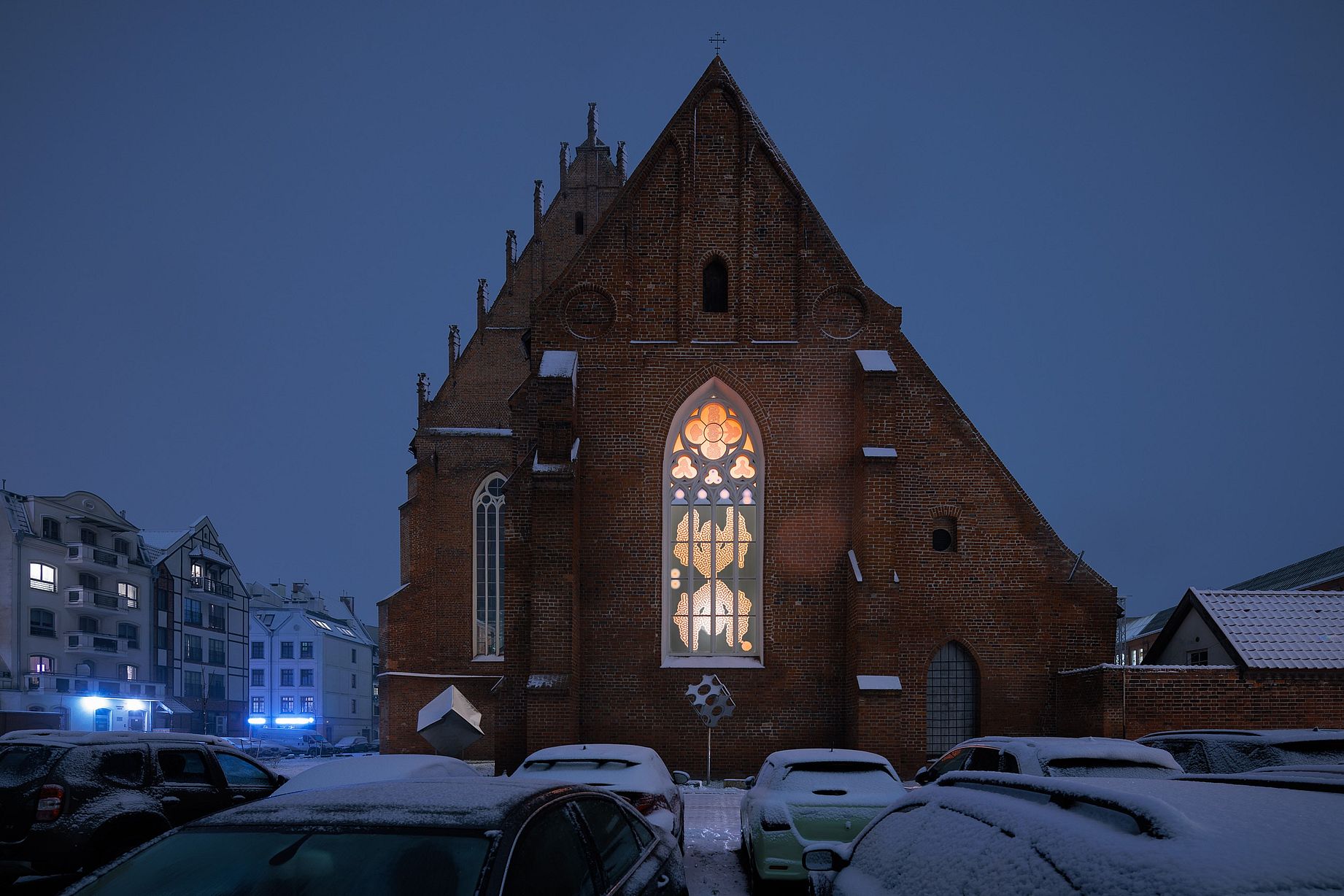 Kościół, w oknie witraż - praca artystyczna, zdjęcie po zmroku, klimat wieczoru, przed kościołem parking, zaśnieżone auta