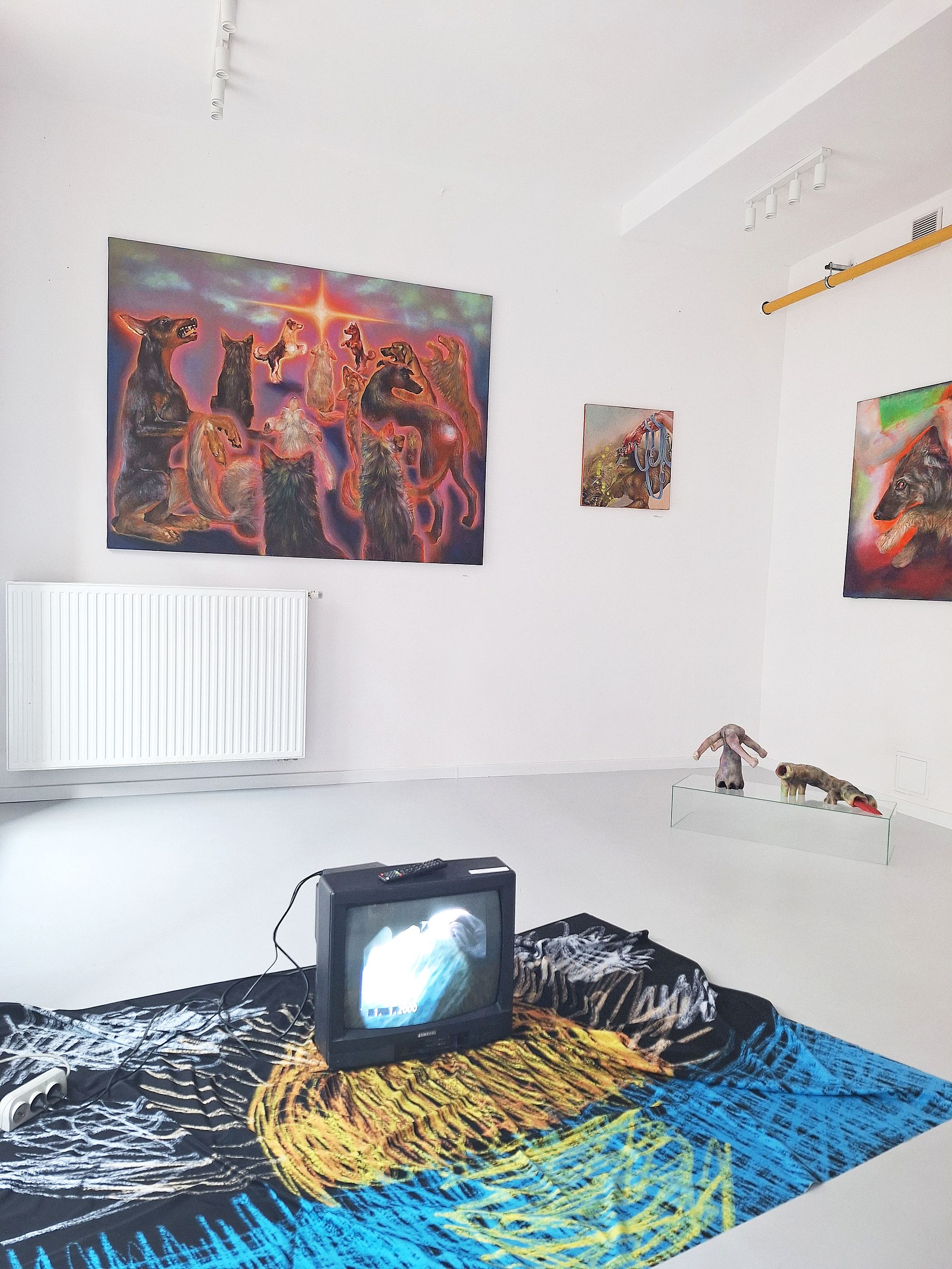 Wystawa w galerii, na ścianie kolorowe obrazy ze zwierzetami, na podłodze instalacja z video na małym monitorze stojącym na kolorowej narzucie