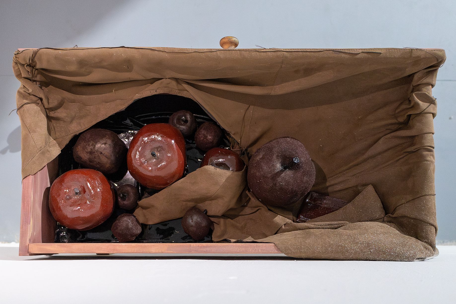 Na zdjęciu martwa natura w formie rzeźby z tkaniny lub wosku, ukazującą zgniłe jabłka ułożone w drewnianej skrzynce i częściowo przykryte brązowym materiałem.