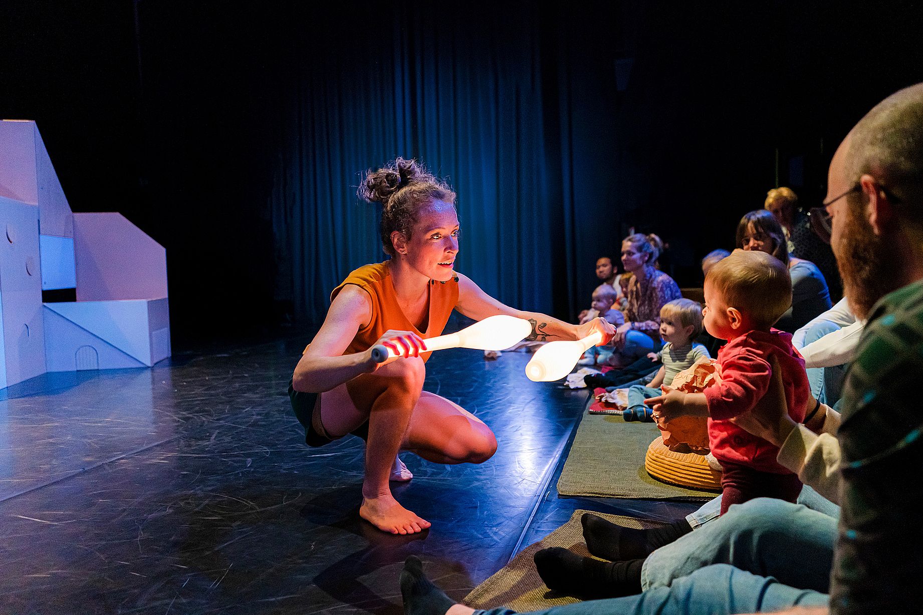 Na zdjęciu performerka w pomarańczowej koszulce i krótkich spodenkach, trzymająca świecące maczugi do żonglerki, kuca na scenie przed grupą małych dzieci i rodziców, nawiązując z nimi bezpośredni kontakt.