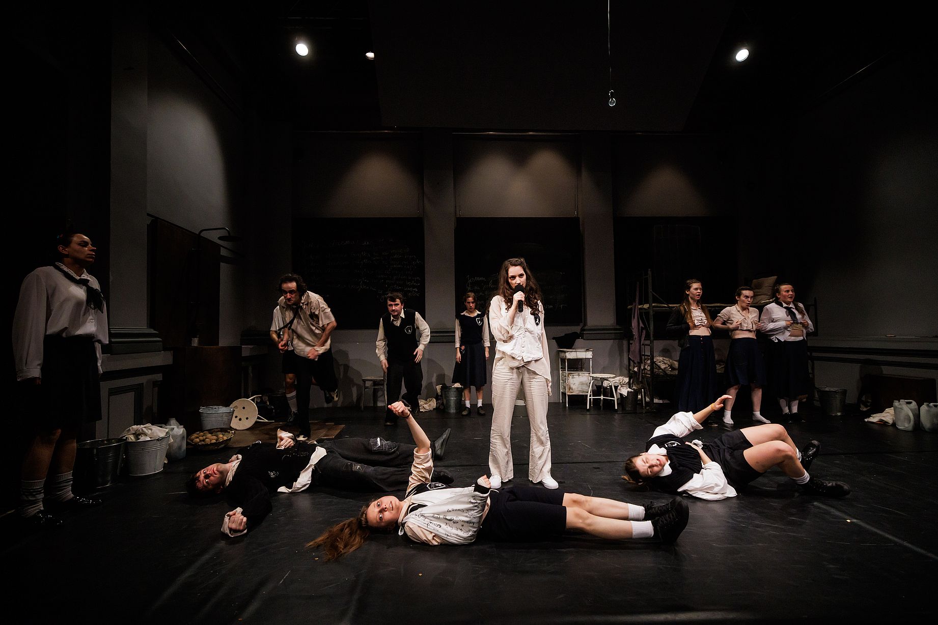 scena w teatrze, aktorka w centralnym punkcie mówi lub śpiewa do mikrofonu, część aktorów leży