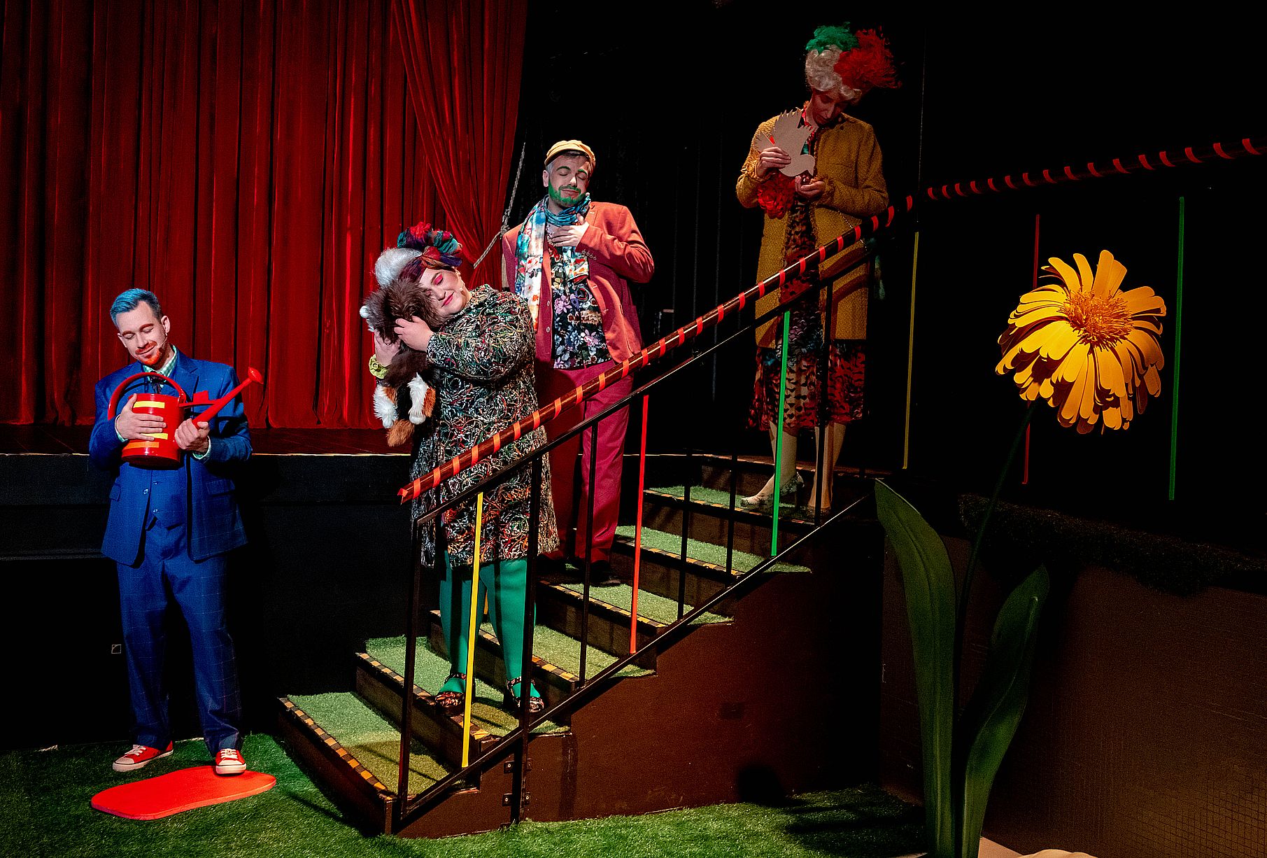 Grupa aktorów w teatrze stoi na schodach w trochę dziwnym strojach, na pierwszym planie duży, żółty kwiat