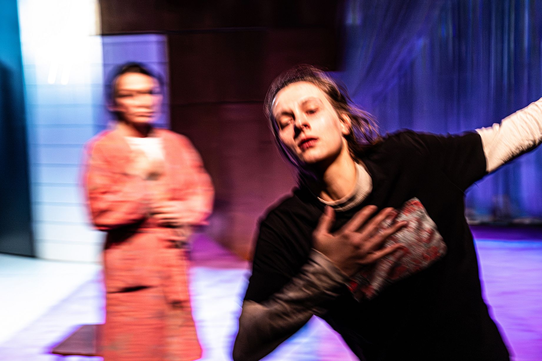 Rozmyte zdjęcie ze spektaklu, osoba w czarnej koszulce w ruchu, z tyłu osoba przygląda się tej scenie