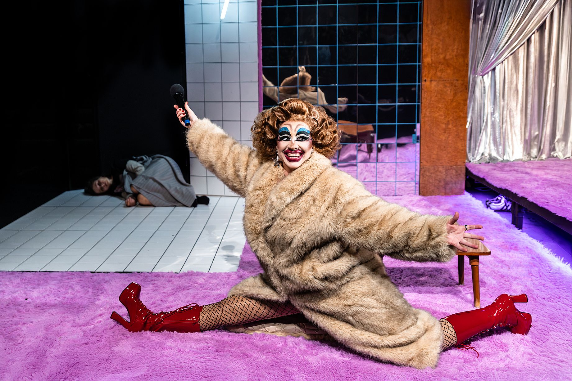 Zdjęcie przedstawia osobę w pełnym makijażu drag queen, ubraną w duży, puszysty płaszcz z futra w jasnym kolorze i czerwone, lakierowane kozaki na wysokim obcasie. Postać siedzi na jasnofioletowym, włochatym dywanie, z szeroko rozłożonymi nogami i ramionami, trzymając w uniesionej ręce mikrofon. Wyraz twarzy jest radosny, z szerokim uśmiechem i mocnym makijażem oczu.