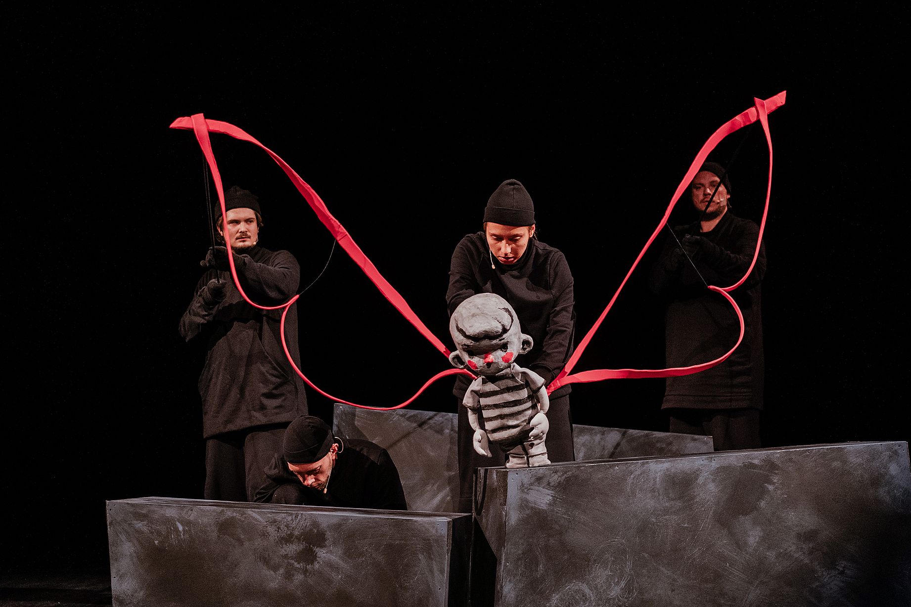 Na scenie w mrocznym oświetleniu trzech ubranych na czarno aktorów animuje dużą lalkę chłopca o szarym ciele i czerwonych policzkach, używając czerwonej wstążki 