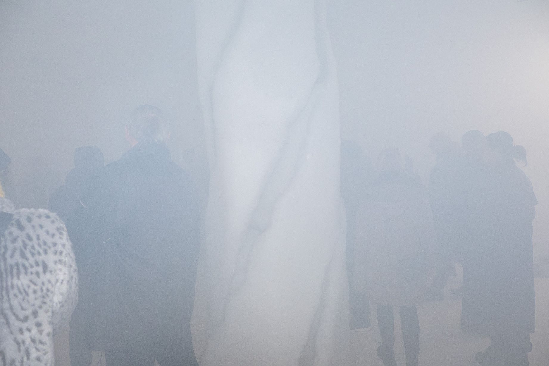 Zdjęcie przedstawia grupę osób stojących w gęstej, białej mgle, z jasnym, pionowym obiektem w centrum. Postacie są słabo widoczne i w większości stanowią sylwetki z powodu słabego oświetlenia i dymu. 