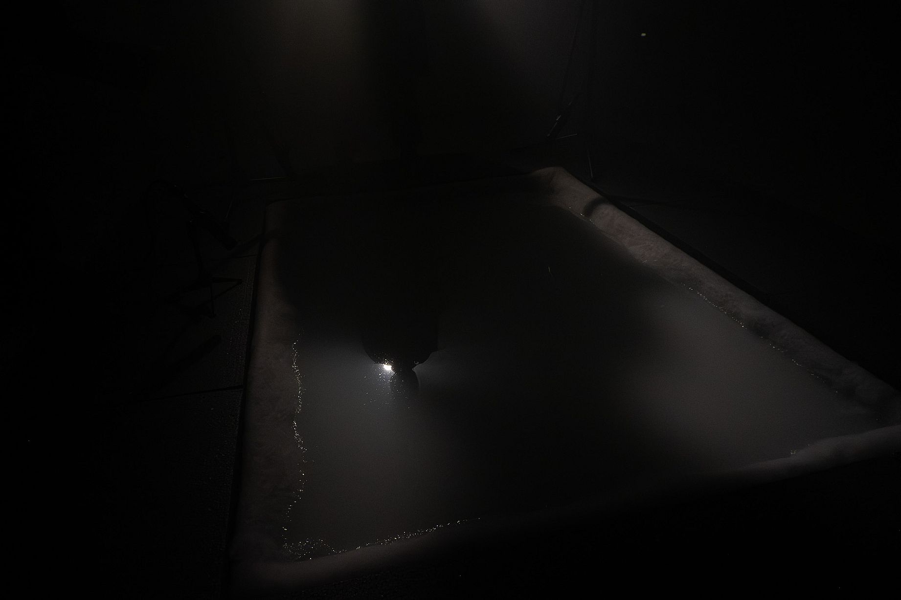 Zdjęcie przedstawia osobę w wannie lub basenie w bardzo słabym oświetleniu, z pojedynczym, jasnym źródłem światła odbijającym się od wody