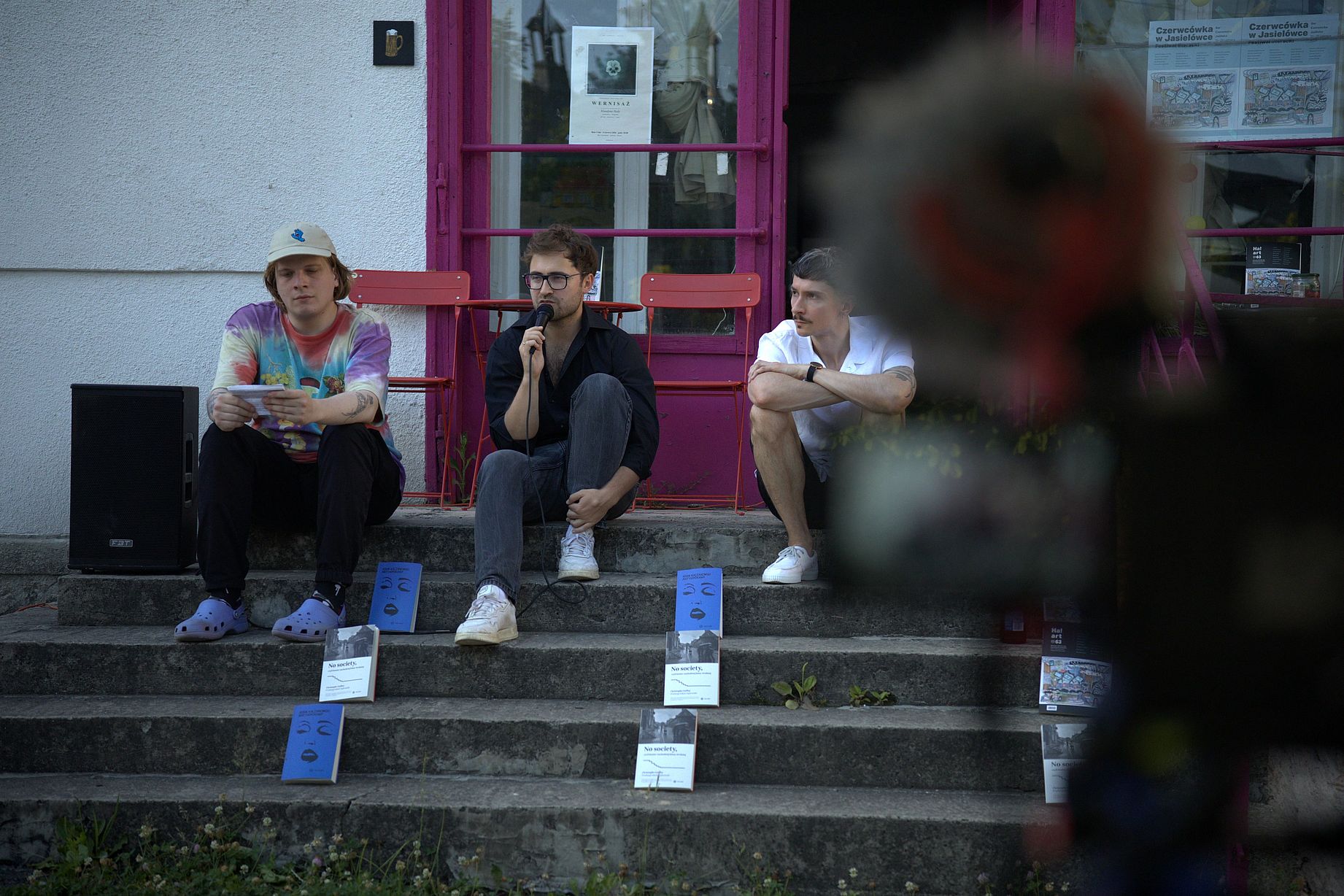 Trzech mężczyzn siedzi na schodach, na których stoi kilka książek, osoba w środku przemawia przez mikrofon