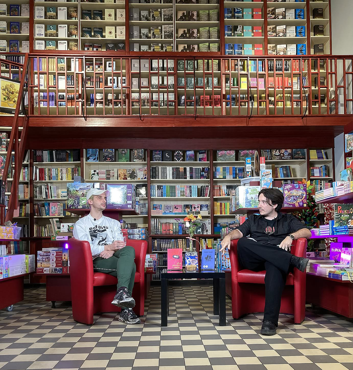 Dwóch ężczyżn na spotkaniu literackim w księgarni. Na podłodze kafelki w kratkę, czerwone meble, dużo książek na półkach