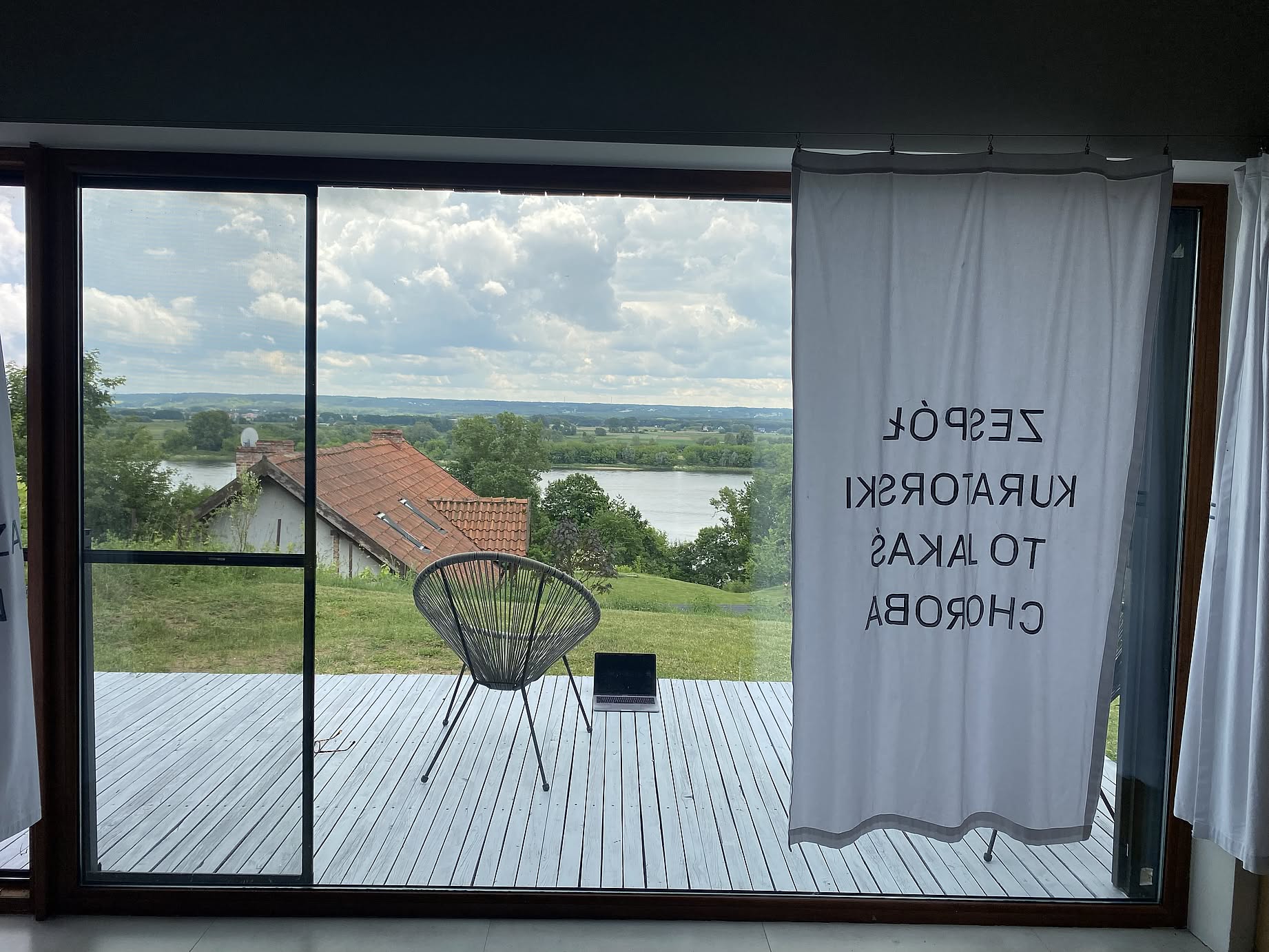 zdjęcie robione z wnętrza domku z dużą szybą, zasłona z napisem: Zespół kuratorski to jakaś choroba, na tarasie krzesło, i laptop, w tle rzeka