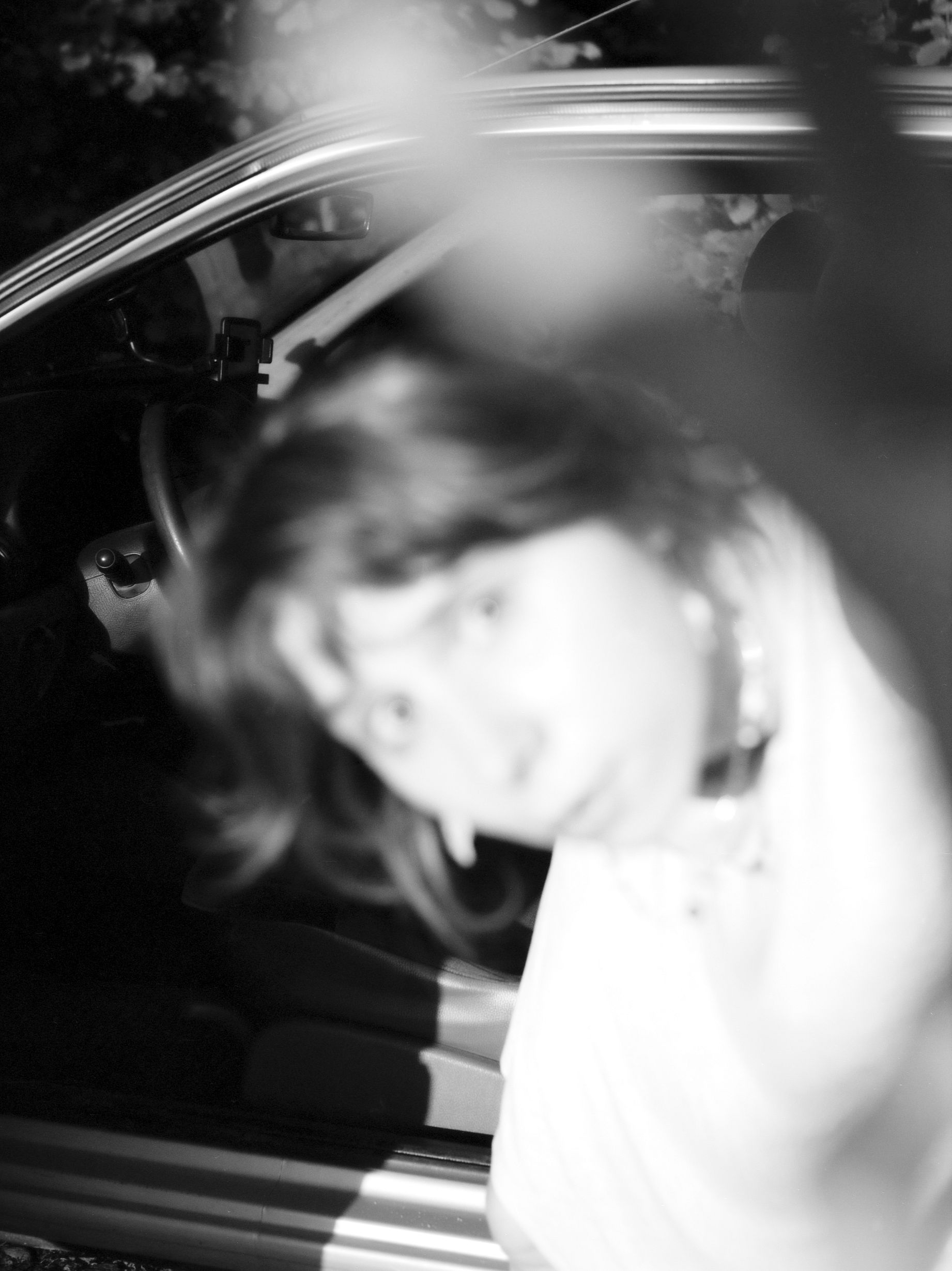 Rozmyte, czarno-białe zdjęcie przedstawia osobę, prawdopodobnie kobietę, wychylającą się z samochodu, z widocznym wnętrzem pojazdu i elementami odbicia światła na pierwszym planie.