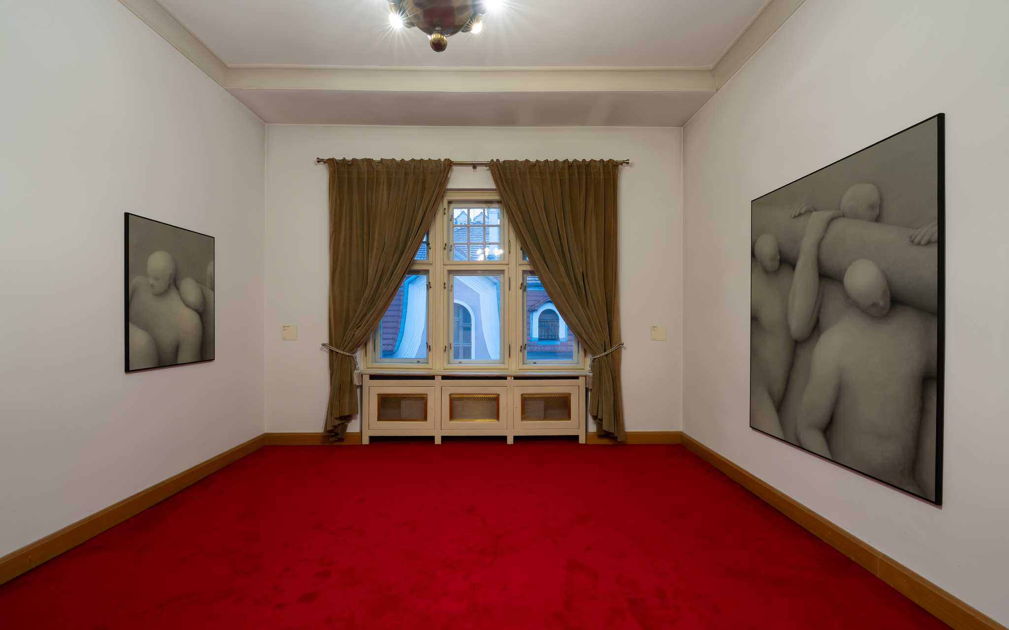 Pomieszczenie galerii lub pokój mieszkalny z białymi ścianami i czerwoną wykładziną, ma okno z brązowymi zasłonami i dwa duże, czarno-białe obrazy przedstawiające nagie postaci wiszące na przeciwległych ścianach.
