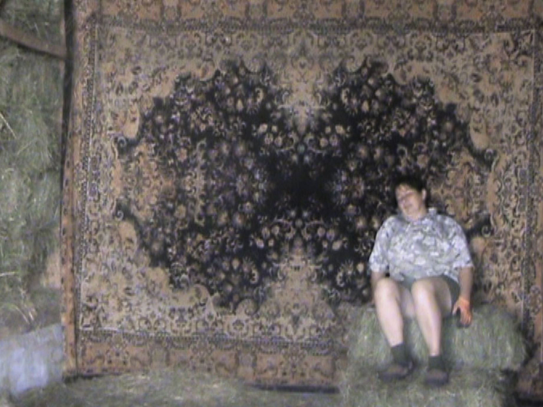 Zdjęcie przedstawia osobę siedzącą na belach siana przed dużym, tradycyjnym dywanem wiszącym na ścianie