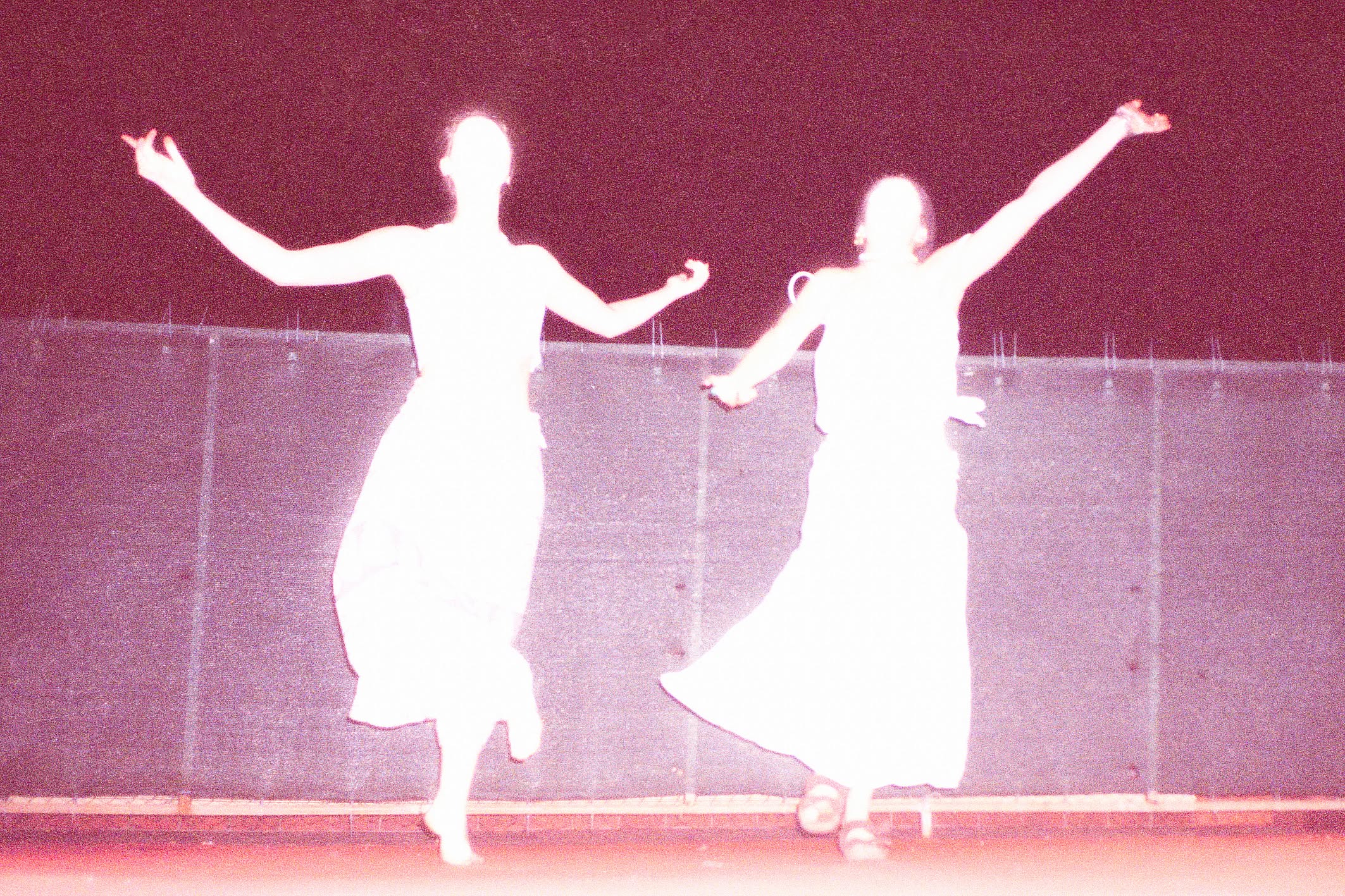 Zdjęcie w odcieniach magenty przedstawia dwie postacie w jasnych, zwiewnych strojach, zastygłe w dynamicznych pozach tanecznych na scenie.