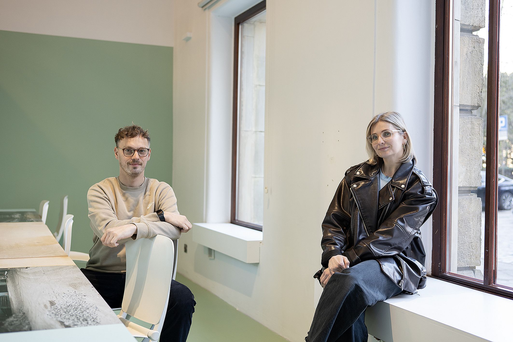Dwie osoby siędza we wnętrzu biura, mężczyzna po lewj jest w okularach, kobieta siedzi na parapecie, obrana w kurtkę ze skóry
