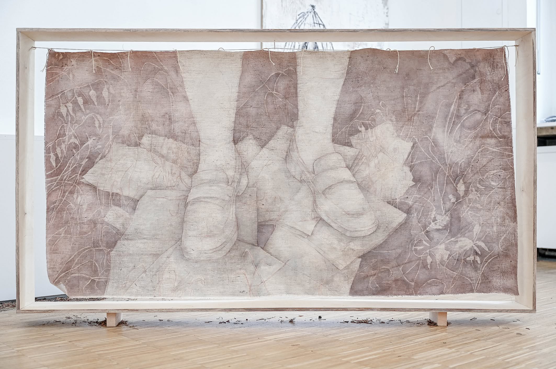 Prezentowana instalacja artystyczna, umieszczona w prostej, drewnianej ramie ekspozycyjnej, przedstawia wielowarstwową kompozycję z tkanin w odcieniach brązu i beżu, zawieszonych na sznurkach, z widocznymi abstrakcyjnymi wzorami przypominającymi korzenie 