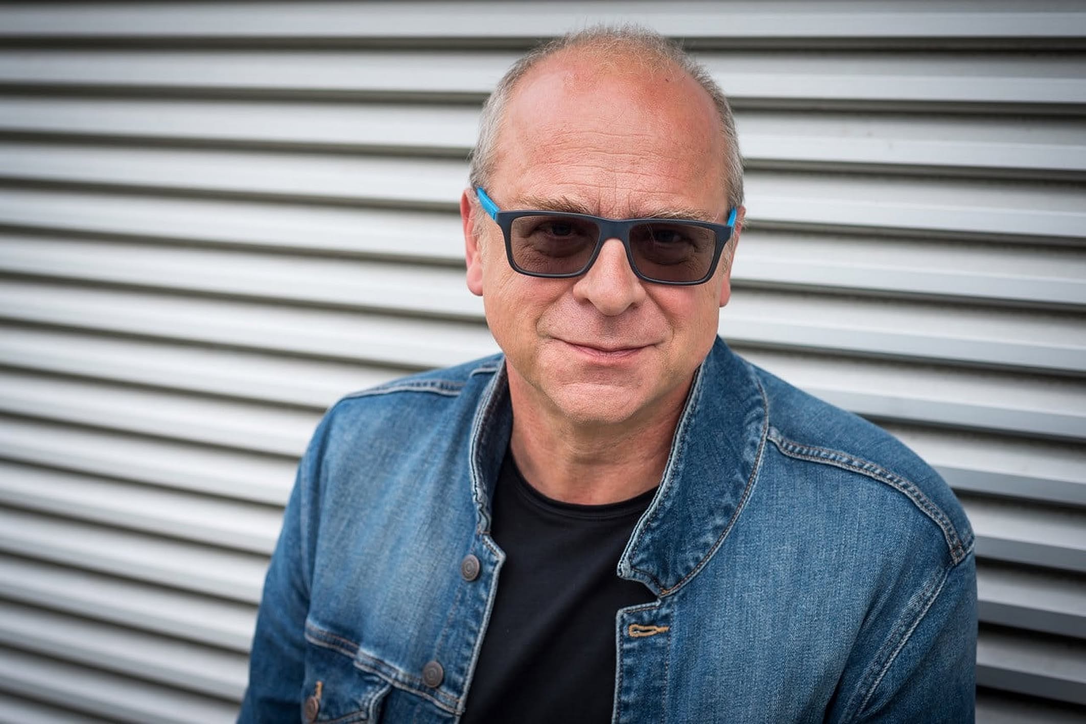 Na zdjęciu znajduje się mężczyzna w średnim wieku, łysiejący, uśmiechnięty, patrzący prosto w obiektyw. Ma na sobie niebieską, dżinsową kurtkę i czarny podkoszulek. Nosi prostokątne okulary przeciwsłoneczne z niebieskimi akcentami na oprawkach. Tło stanow