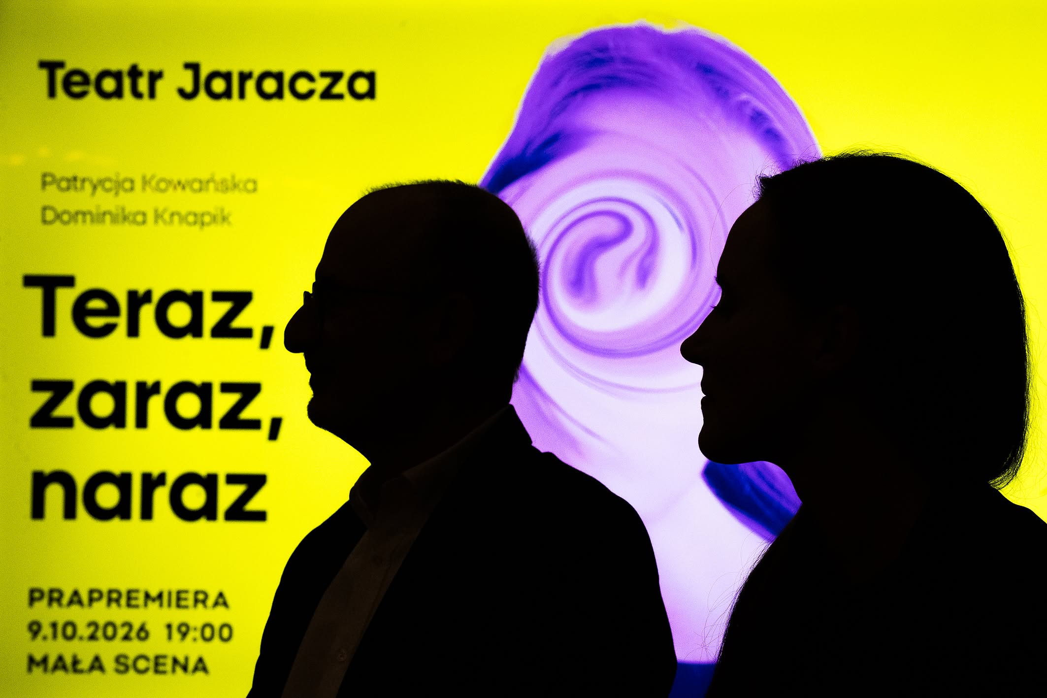 Cienie kobiety i mężczyzny, w tle podświetlony  plakat spektaklu Teatru Jaracza, żółte tło