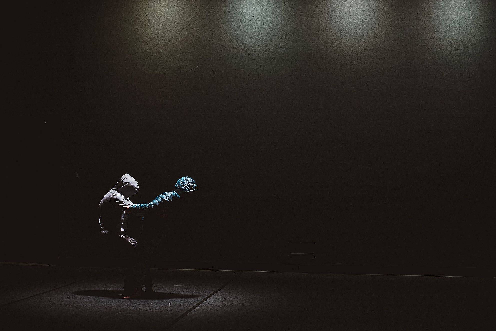 Na ciemnej scenie dwie osoby w kapturach i kurtkach stoją naprzeciw siebie, jedna podtrzymuje drugą, która odchyla się do tyłu w dramatycznym geście.