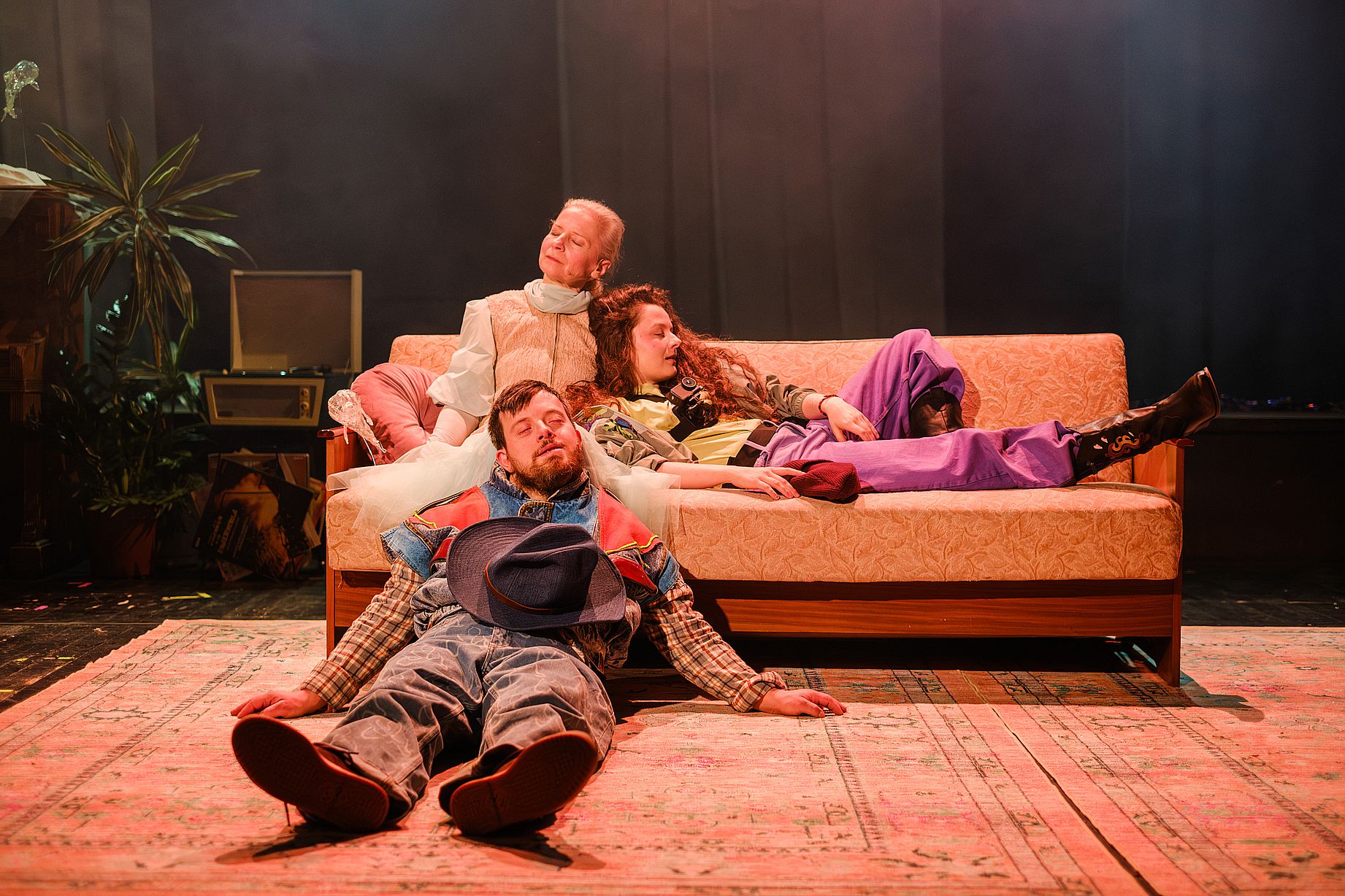 scena w teatrze, dwie aktorki leża na kanapie, aktor z kapeluszem  siedzi na ziemi i opiera się o kanapę