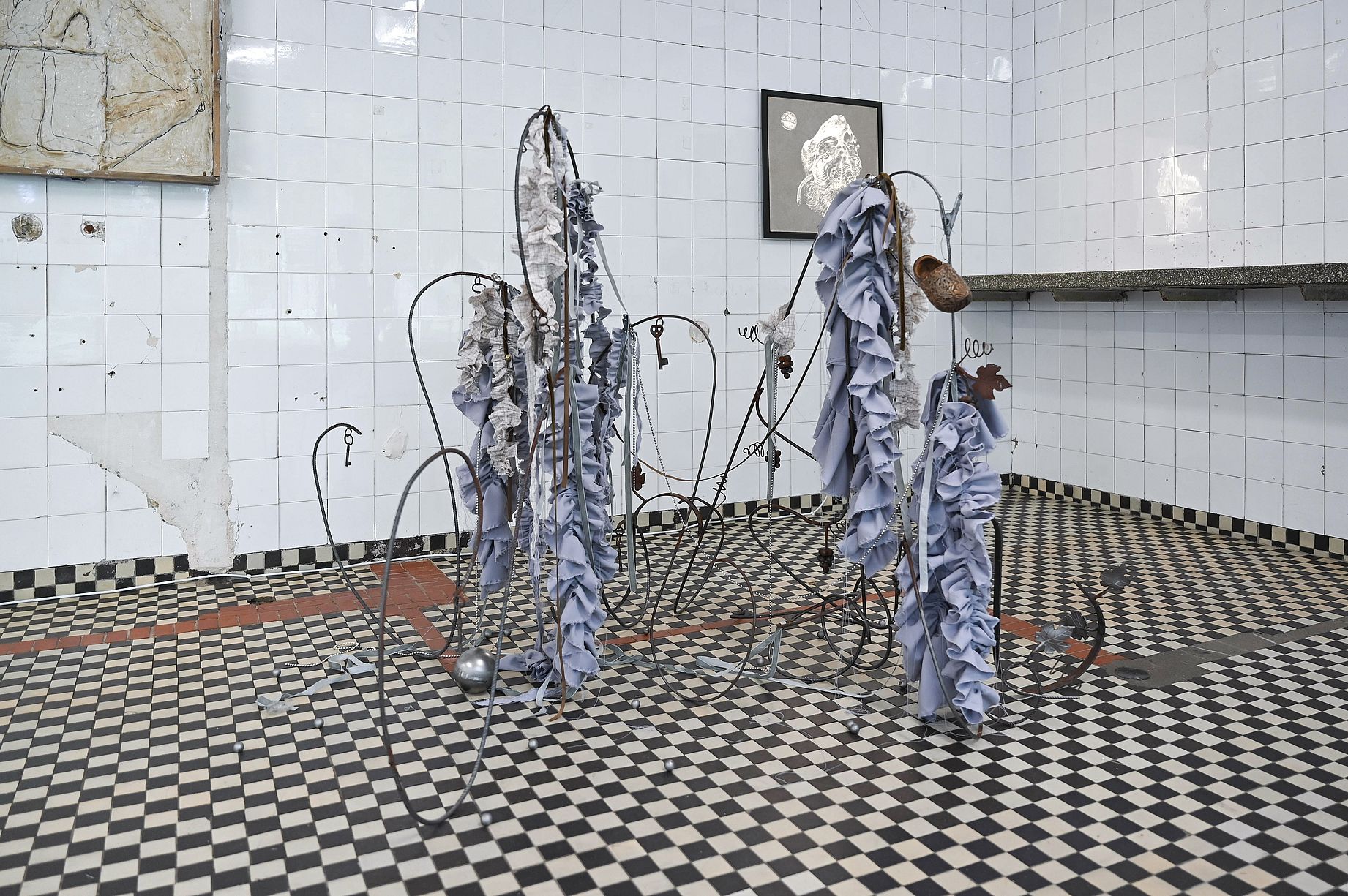 Na zdjęciu widać instalację artystyczną z mieszanych mediów, wykonaną z pogiętych metalowych prętów i zwisających, niebieskoszarych tkanin, umieszczoną w pomieszczeniu wyłożonym biało-czarnymi kafelkami, prawdopodobnie podczas wystawy w galerii