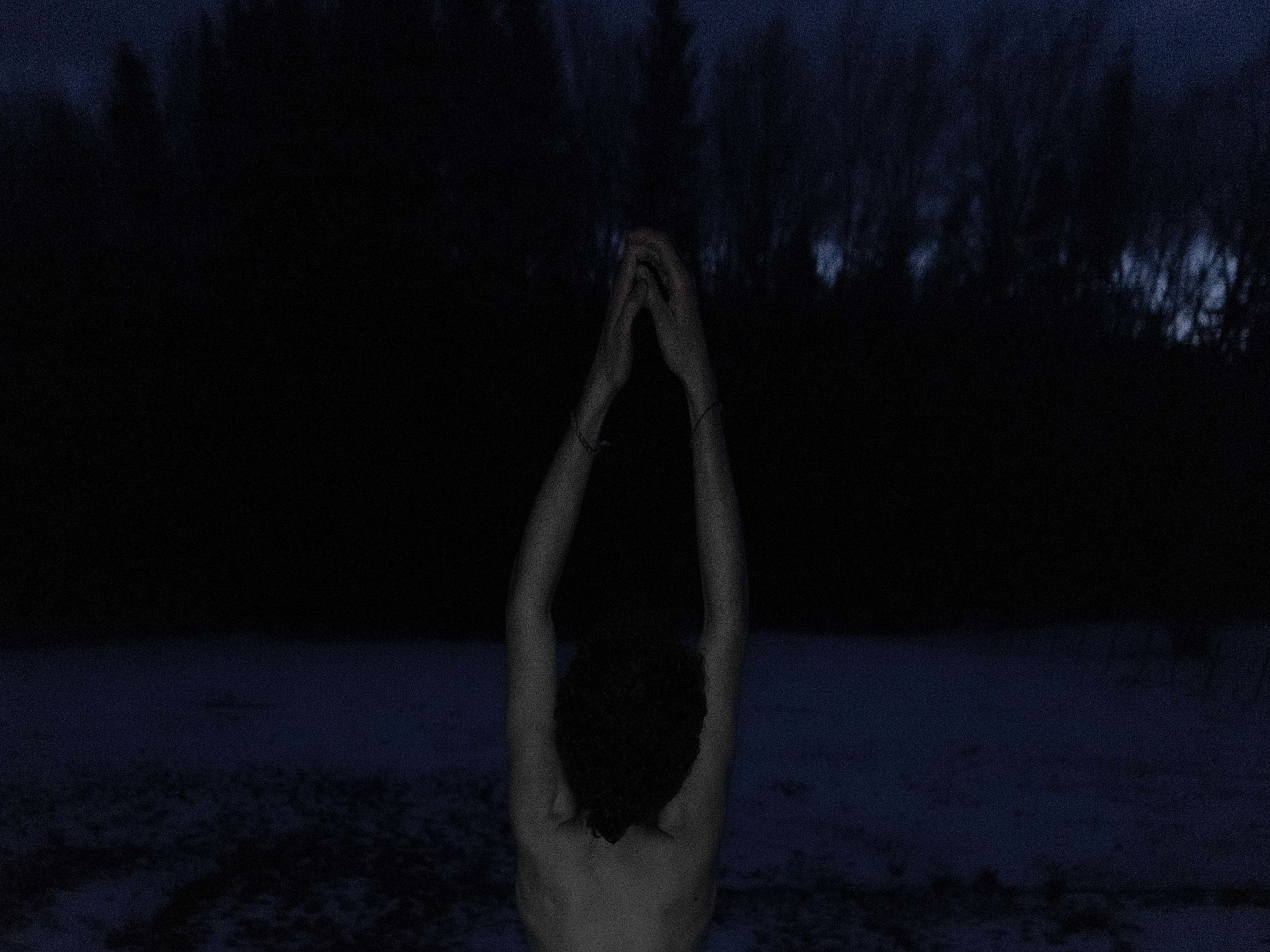 Zdjęcie przedstawia osobę stojącą tyłem do obiektywu w ciemnym otoczeniu, prawdopodobnie w nocy, z rękami uniesionymi nad głową i złączonymi, co sugeruje pozycję jogi lub medytacji.