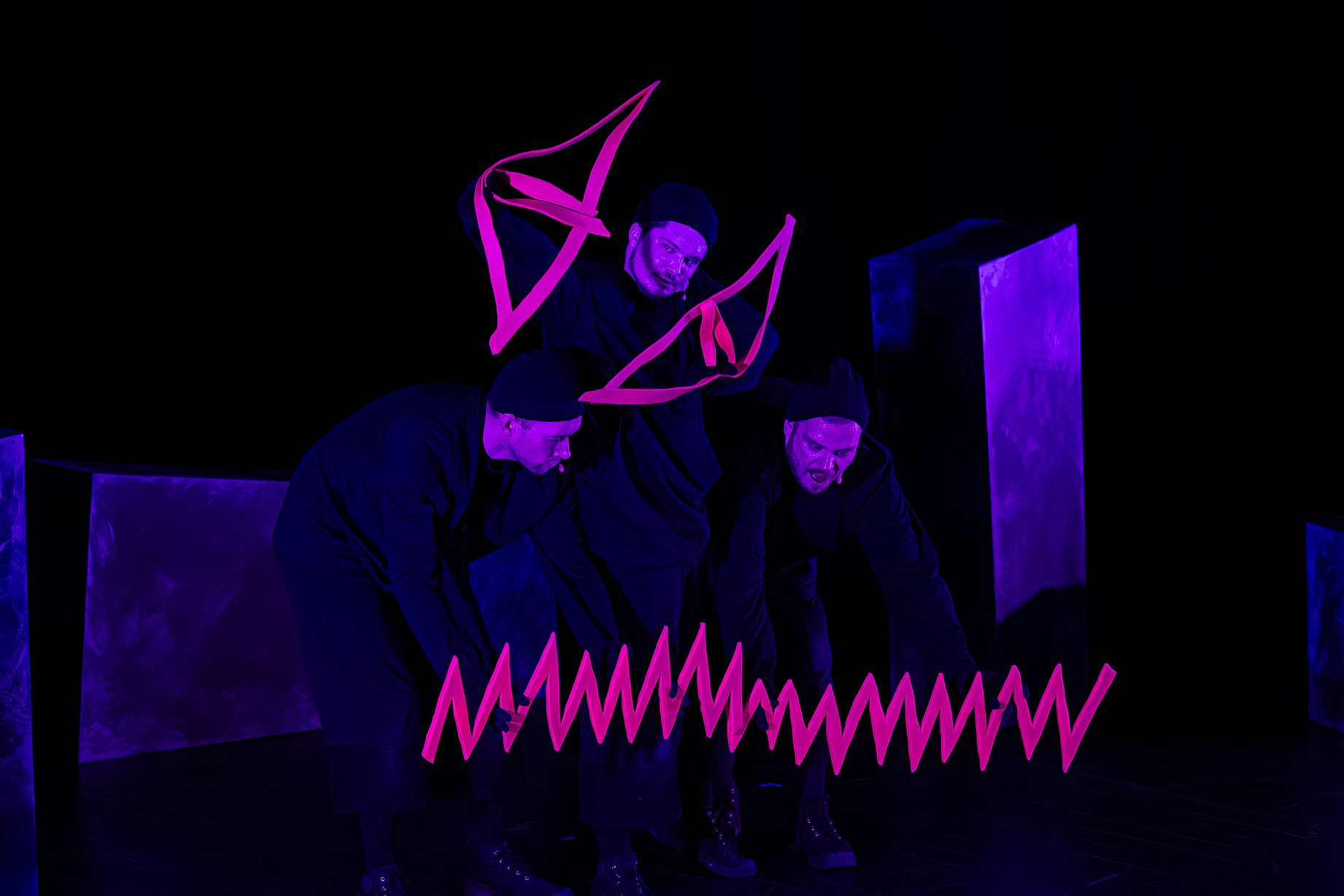 Na scenie w mrocznym oświetleniu trzech ubranych na czarno aktorów animuje używając czerwonej wstążki, podświetlonej fluorescencyjnie 